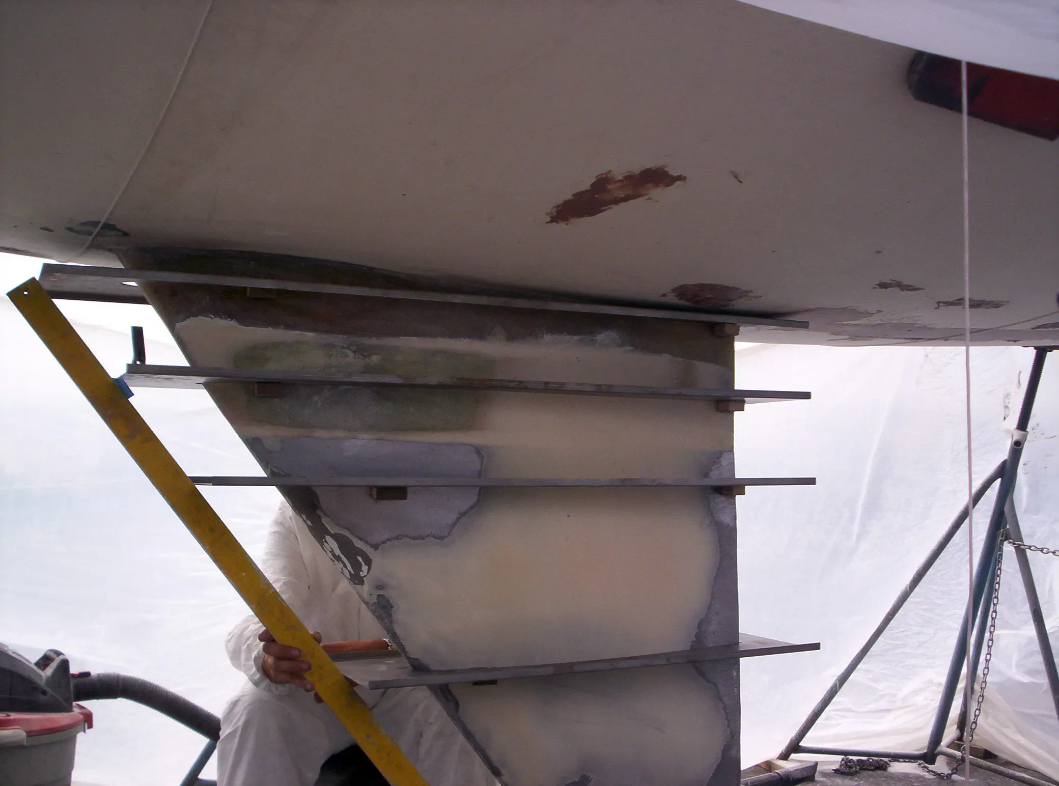 A partially constructed boat hull with support beams and a person's hand holding a measuring tool, set in a work area covered with plastic.