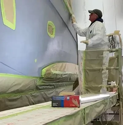 A painter in a protective suit applies paint to a boat's hull, surrounded by drop cloths and tape, in a well-lit workshop.