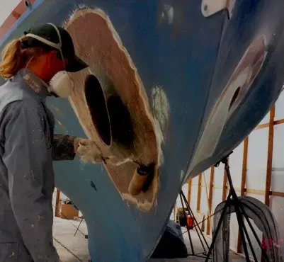 A worker in a mask repairs a boat's hull, sanding an area with two large cutouts while surrounded by wooden scaffolding.