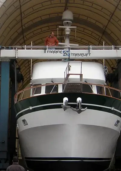 A large boat is being lifted by a Marine Travelift, with a worker overseeing the process from the lift's upper platform.
