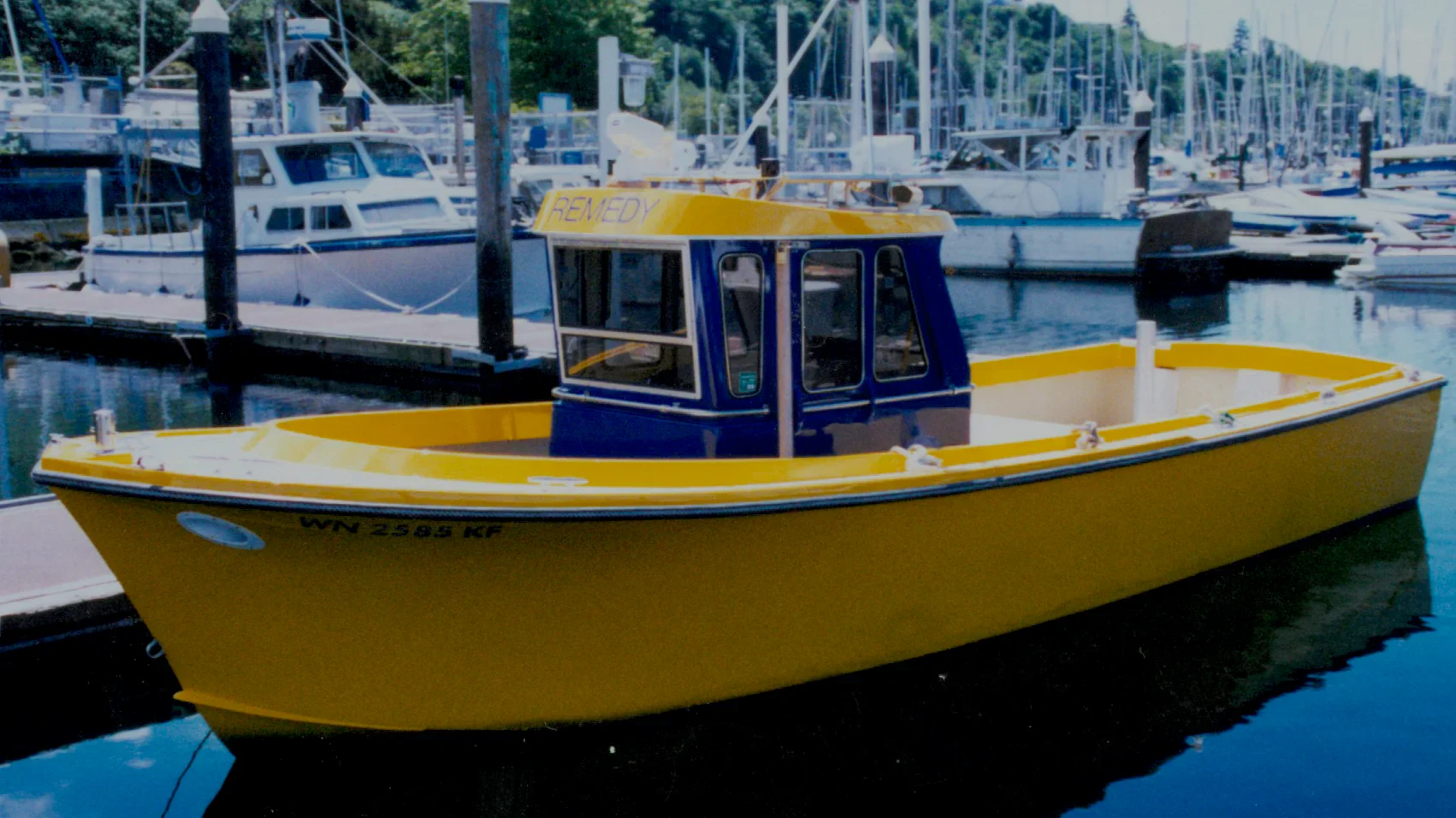 A bright yellow boat with a blue cabin docked in a marina, surrounded by other boats and masts reflected in the water.
