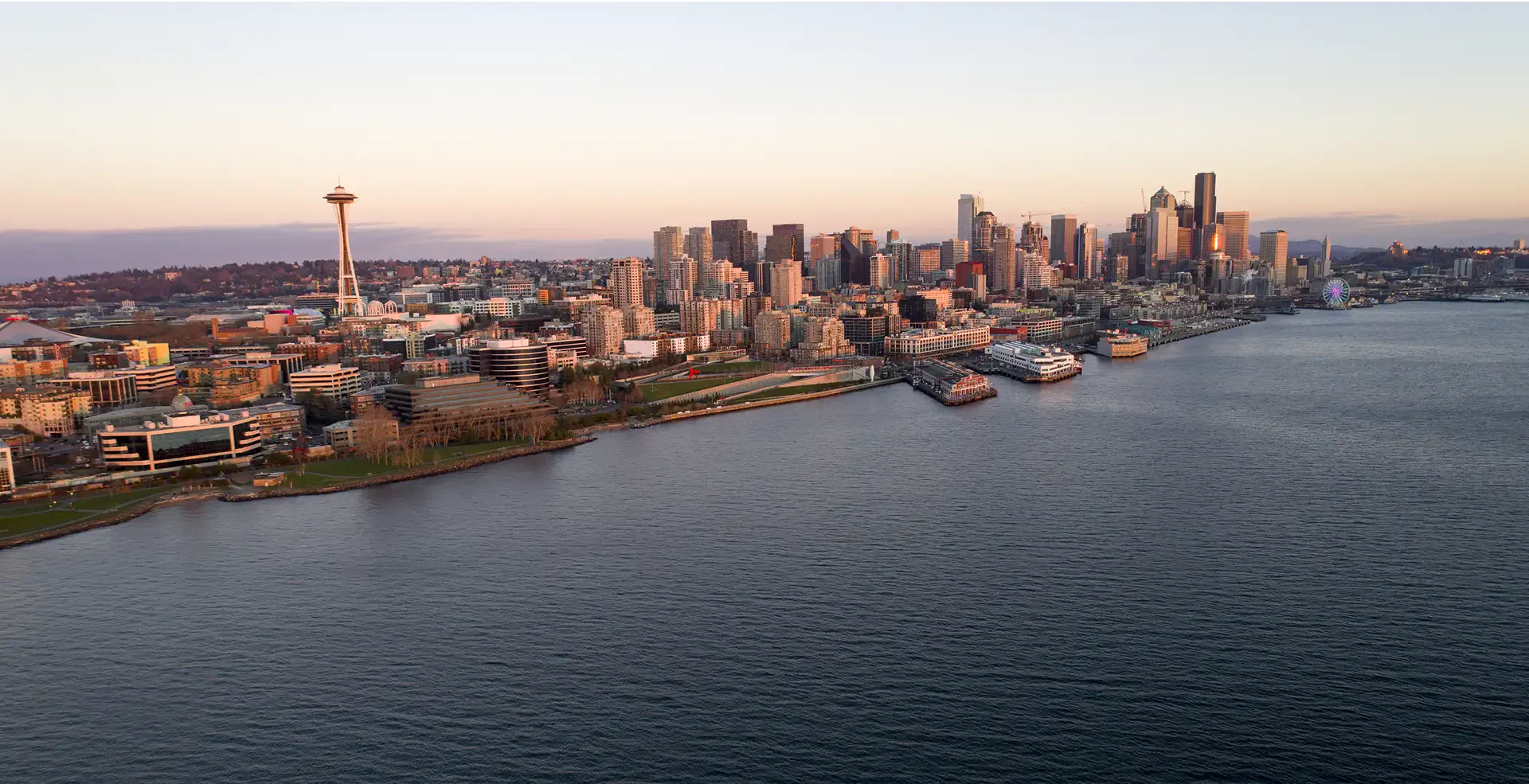 Aerial view of Seattle's skyline at sunset, featuring the Space Needle, waterfront, and cityscape reflecting in the water.