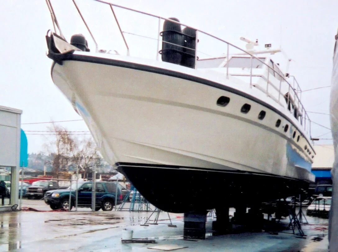A large white boat on stilts in a marina for maintenance, surrounded by vehicles and buildings under a cloudy sky.
