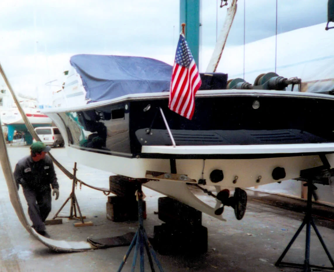A technician works on a boat on stands in a marina, featuring an American flag on the bow and a cloudy sky overhead.